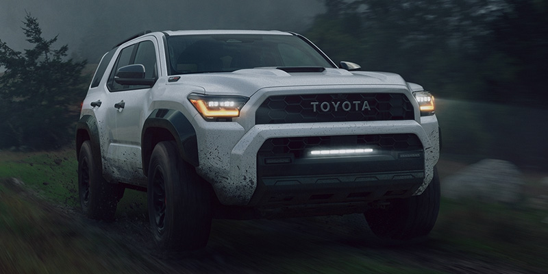 A white 2025 Toyota 4Runner TRD Pro driving on a muddy trail in a misty forest, showcasing its rugged off-road design with illuminated headlights and a light bar.
