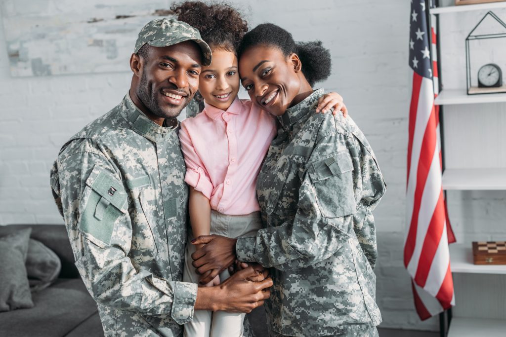 Two adults in military uniforms smiling and embracing a young child indoors, with an American flag visible in the background.