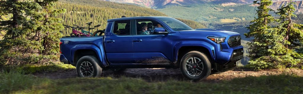 Blue 2026 Toyota Tacoma pickup truck driving off-road through a forested mountain trail with bikes in the bed, highlighting its rugged adventure capability.