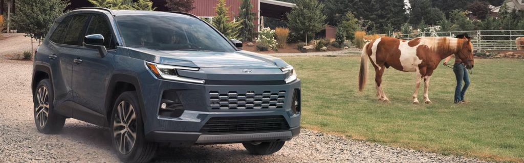 Blue 2026 Toyota RAV4 SUV driving on a rural gravel road near a farm, highlighting its versatile design and everyday utility for Denison drivers.