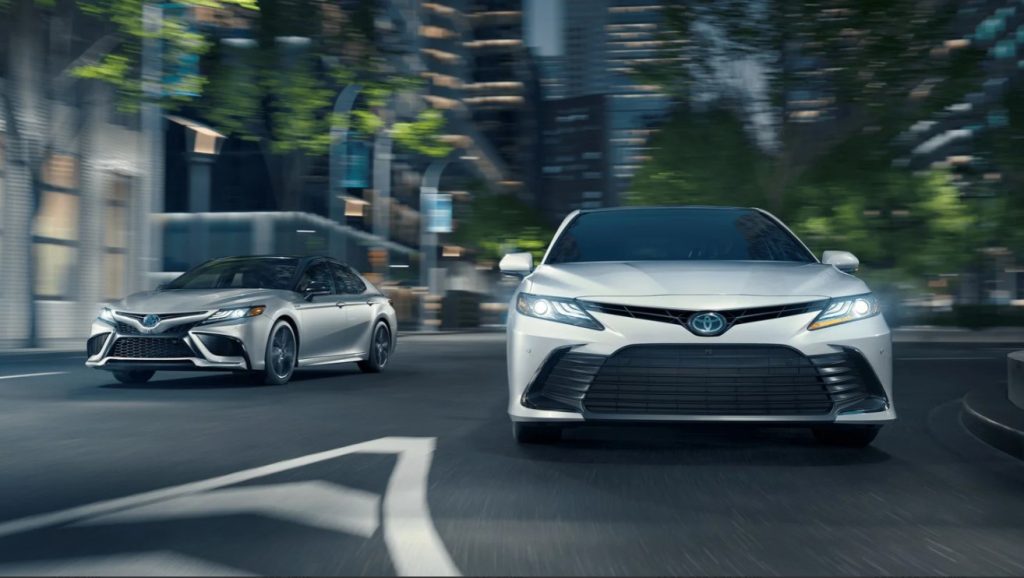 Two Toyota sedans driving on a city street at speed, shown in a dynamic front-facing motion blur scene with modern buildings and trees in the background.