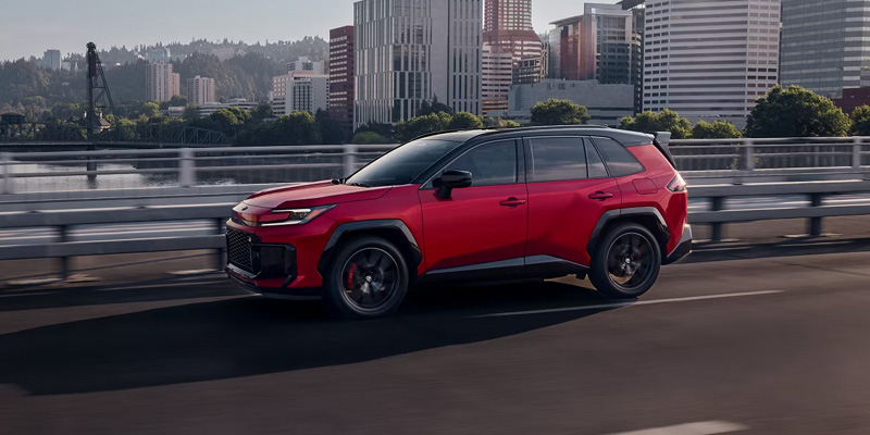 Red Toyota SUV driving on a city bridge with an urban skyline in the background.