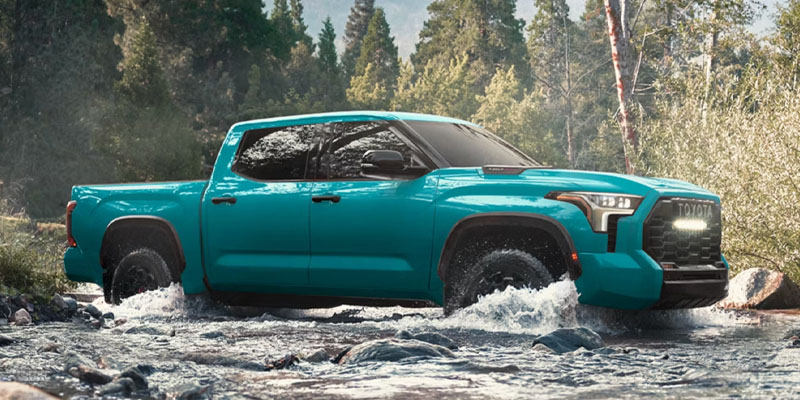 Blue Toyota Tundra truck driving through a shallow river surrounded by trees.