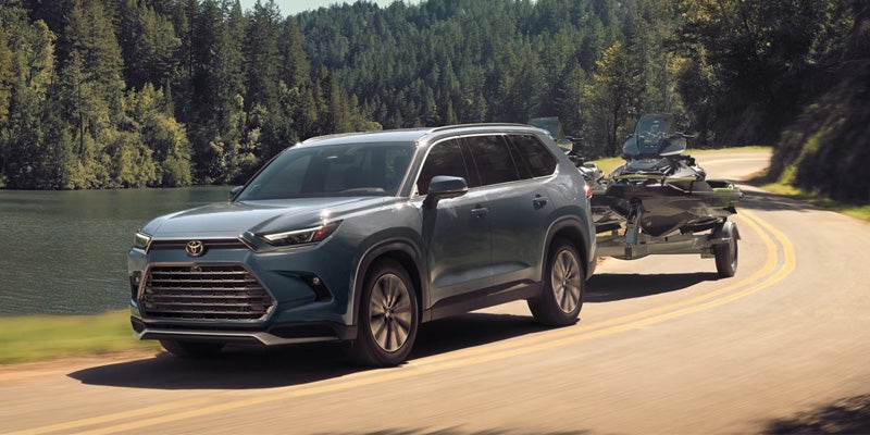 Gray Toyota Grand Highlander SUV towing a personal watercraft trailer on a forest-lined road.